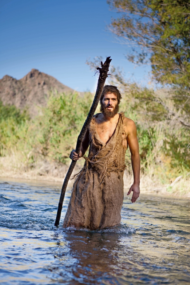 White Male Dressed as Jesus in Jewish Attire. Others as John the Baptist/ Prophet with burlap clothes and wood staff near river.
